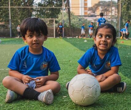 Two children pose sitting next to a soccer ball with their uniforms on, one child looks at the camera and one looks away. 